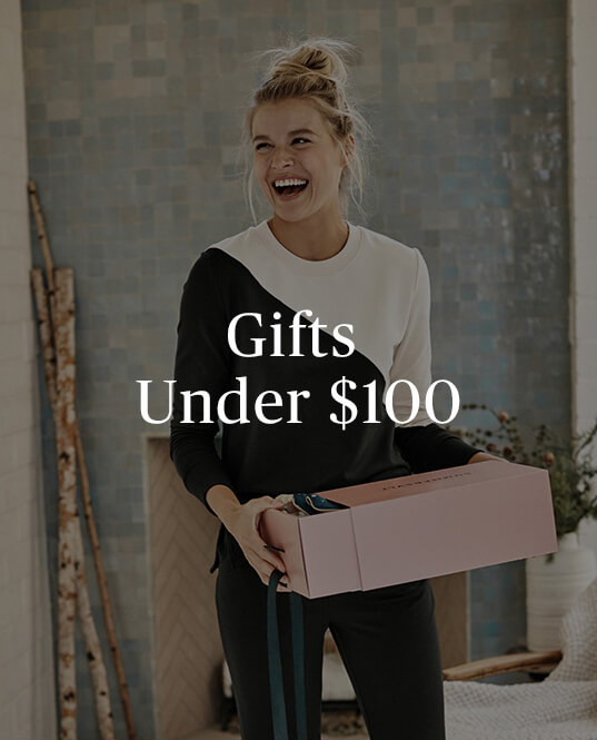 Gifts under $100 copy over an image of a smiling blonde woman holding a pink present wearing the Summersalt Softest French Terry set in Sea Urchin and White Sand