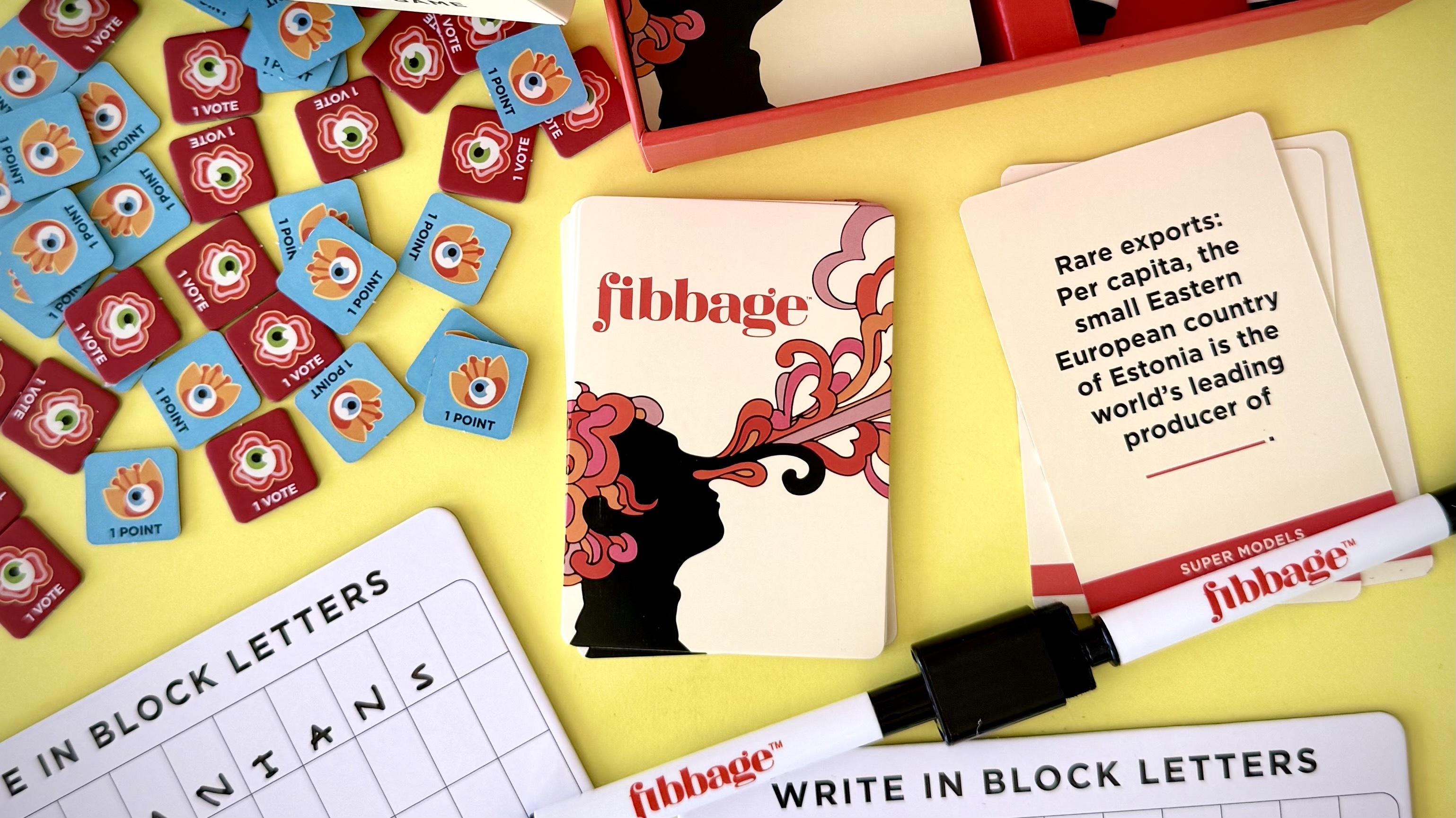 A photo of a Fibbage game card with the Fibbage logo, surrounded by tolkens, the game box, question cards, and white erase board answer cards.