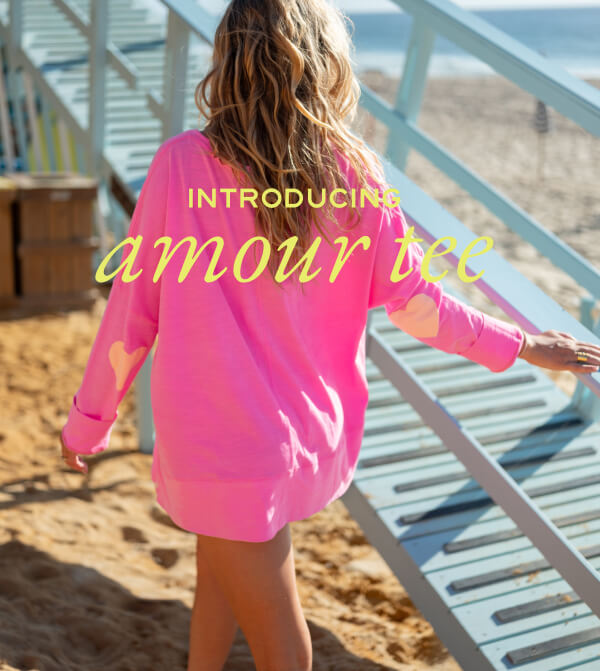 Introducing amour tee - Image of a woman on a beach wearing a long sleeve pink tee with light pink heart patches on the elbows