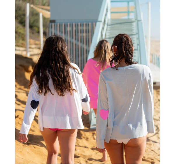 Image of women on the beach wearing different color long sleeve tees with heart patches on the elbows