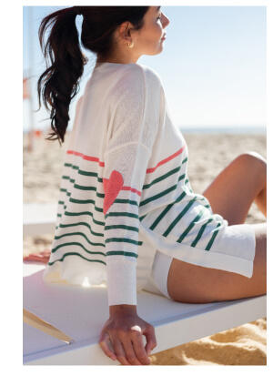 Image of a woman lounging on a beach wearing a white sweater with peach and green stripes and heart patches on the elbows.