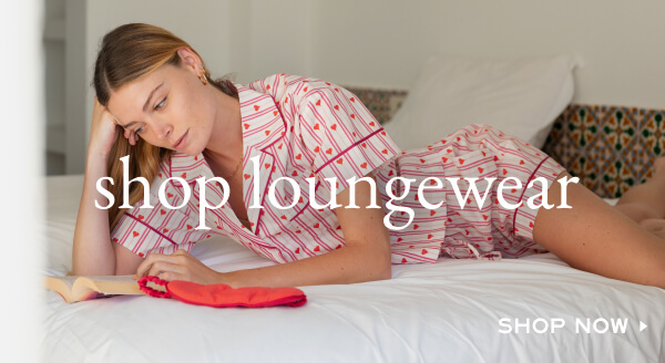 Shop Loungewear - Shop now - Image of a woman lying on a bed, reading and wearing a pajama set with pink stripes and hearts