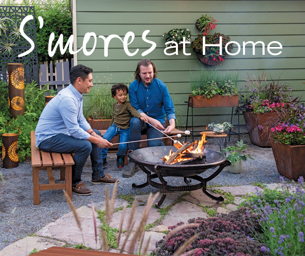 S'mores at Home! Pictured: Young couple with child roasting s'mores over a firepit, surrounded by cylindrical metal lanterns and flower pots full of blooms.