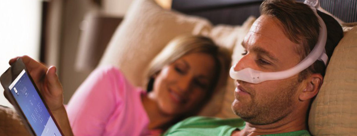 Couple lying in bed. Man using tablet device while wearing CPAP mask.