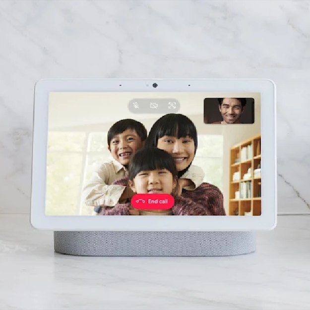 A chalk white colored Google Nest Hub Max is shown on a white marble countertop against a matching backsplash. It is being used for a video call, and two children and an adult woman are shown calling in, with the viewer being a man shown in a video window in the corner of the screen.
